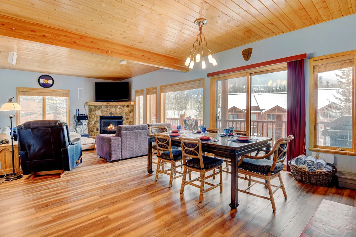 A spacious, sunlit great room with a beautifully set dining table, a prominent gas fireplace, large windows revealing river and ski mountain vistas, and a plush seating area for families to relax together.