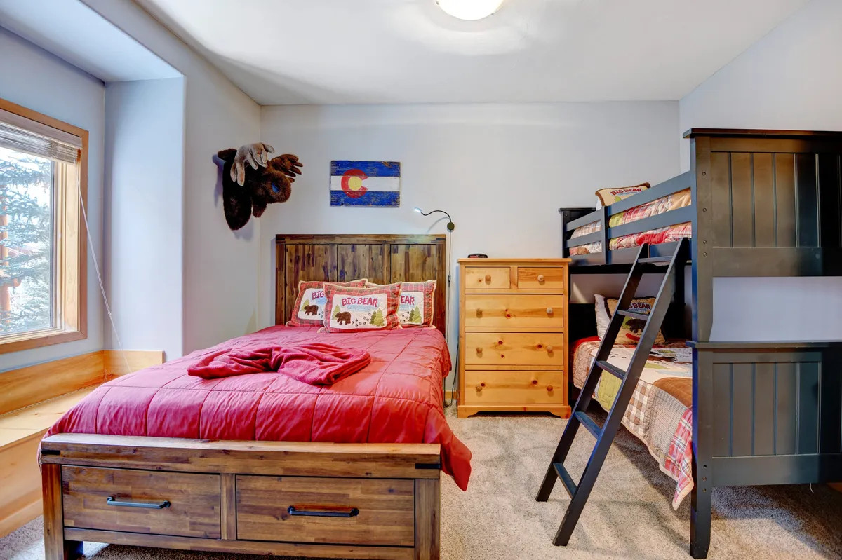 A welcoming guest bedroom displaying a wooden bed with red bedding, a bunk bed suitable for kids or friends, colorful artwork, and a wooden dresser. The room feels cheerful and family-friendly.