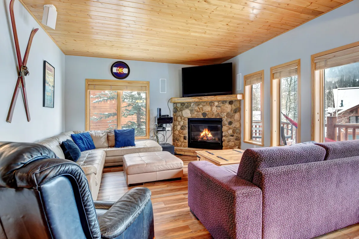A warm living space centered around a gas fireplace, with comfortable seating, a wooden ceiling, and expansive windows framing mountain scenery—the heart of cozy evenings and family movie nights.