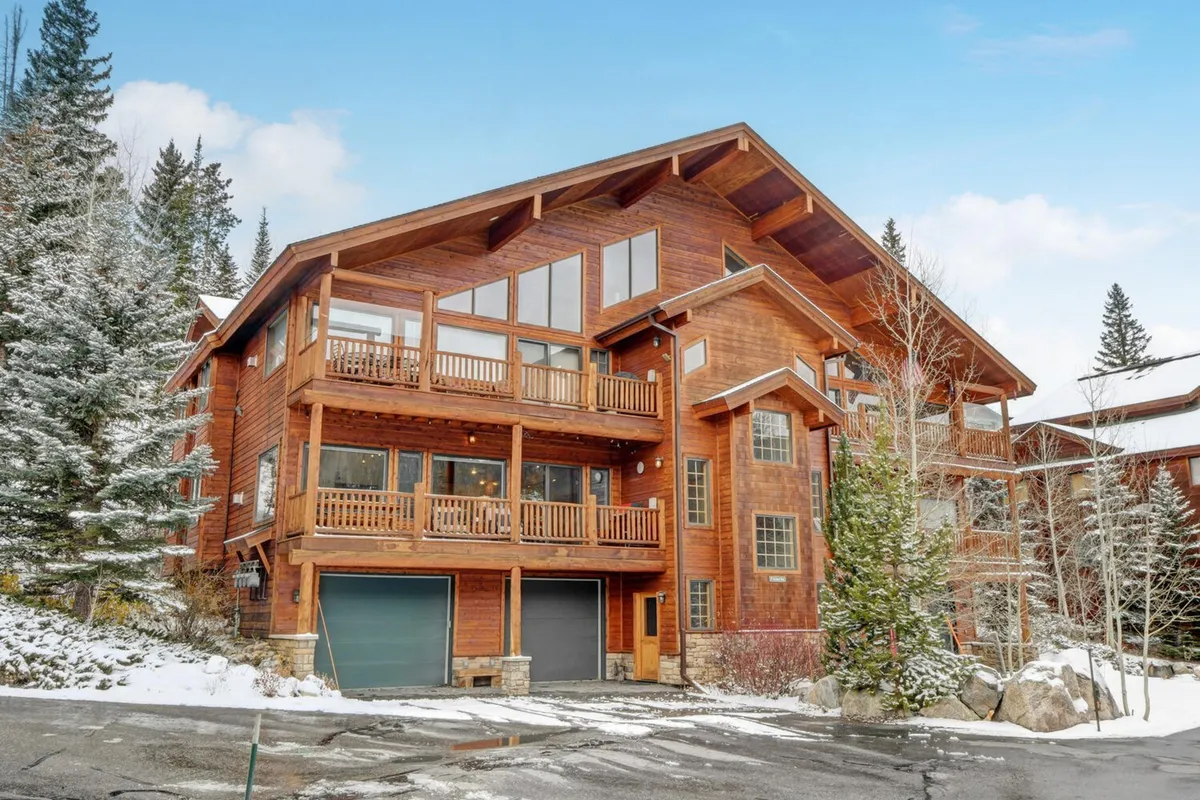 The image shows a welcoming wooden chalet nestled among snow-covered trees, highlighted by large windows and inviting balconies overlooking a serene winter landscape. The cozy exterior glows against the snowy backdrop, suggesting warmth and relaxation, with plenty of space for gathering after adventurous days on the slopes.