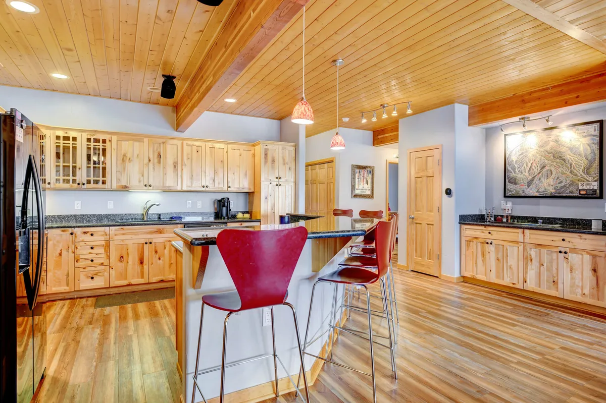 A wide view of the kitchen highlights a large island with comfortable bar stools, wooden cabinetry, granite countertops, and pendant lighting. The welcoming atmosphere suggests a space made for family gatherings, recipe sharing, and milestone celebrations.