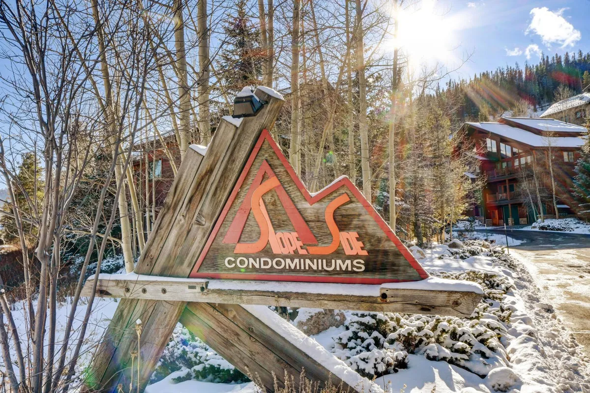 An exterior image showing the 'Slope Side Condominiums' sign, surrounded by trees and a snow-blanketed landscape. The scene conveys a welcoming community atmosphere and hints at nearby parks and recreation areas.