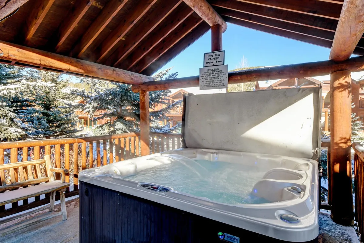 An exterior setting with a communal hot tub surrounded by wooden railings and trees. Guests are enjoying the inviting water, perhaps with a glass of wine, as the snow-covered landscape or warm sunset creates a serene backdrop.